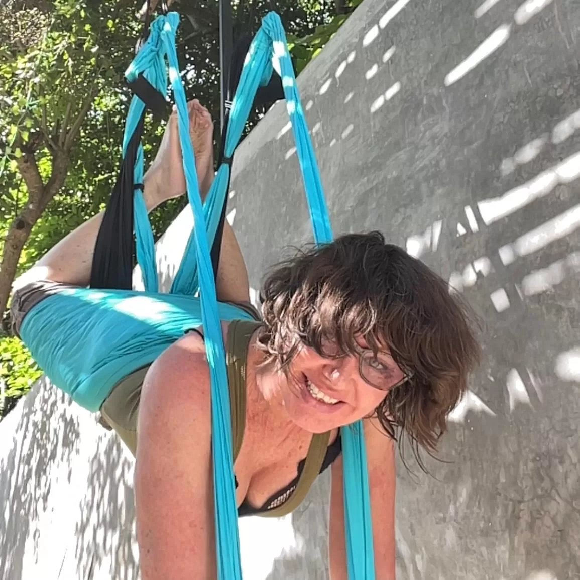 A woman with glasses performs aerial yoga in a blue hammock outdoors, with a concrete wall and trees in the background.